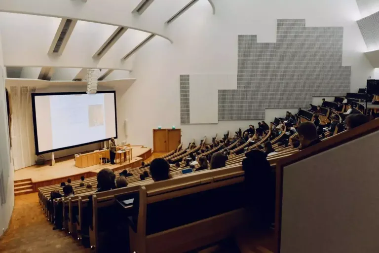 A large, auditorium-style lecture hall, with an instructor lecturing, a slide on a projection screen, and students listening